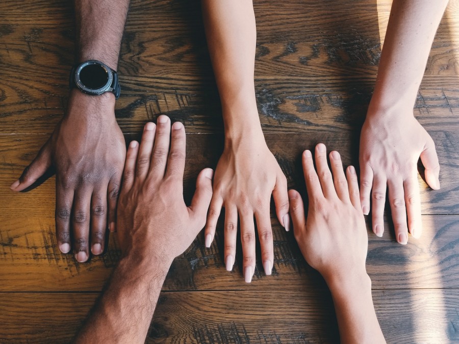 Frauen und Männern haben ihre Hände unterschiedlicher Hautfarben auf einen Holztisch gelegt. Stichwörter: Integration, Ausländeramt, Miteinander, Migration - Photo by Clay Banks on Unsplash