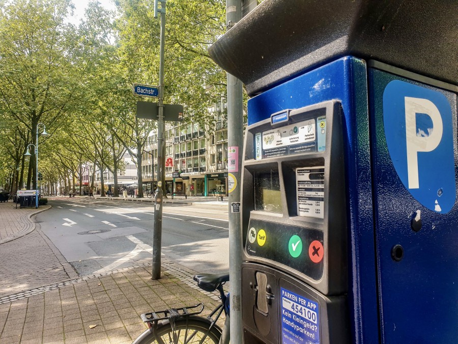 Parkscheinautomat auf der Leineweberstraße, an der Abbiegung zur Bachstraße, mit dem Straßenschild Bachstr., grünen Bäumen und blauem Himmel. Hinter dem Automat lehnt ein Fahrrad. - Onlineteam