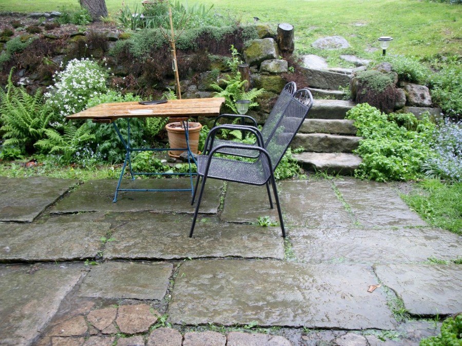 Blick durch den Regen auf eine Terrasse mit Tisch und Stühlen, dahinter geht es auf eine große Gartenwiese. Niederschlagswasser, Versickerung - Canva
