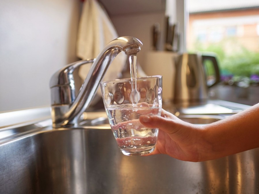 Hand hält ein Glas unter einen fließenden Wasserhahn und füllt es mit Trinkwasser. Legionellen, Wasserverschmutzung, Krankheitserreger, Wasserschutz - Canva