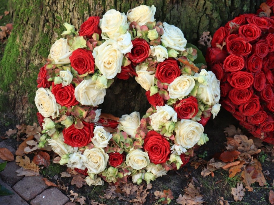 Trauerkranz mit weißen und roten Rosen und Trauerkranz in Herzform aus roten Rosen lehnen an einem Baumstamm. Informationen zum Verhalten im Trauerfall. Friedhof, Friedhöfe, Beerdigungen, Bestattungen, - Canva