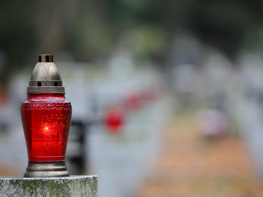 Friedhof, Friedhöfe, Brandgefahr: Das Foto zeigt im Vordergrund ein rotes Grablicht auf einem Steinsockel, der Hintergrund ist verschwommen - Canva