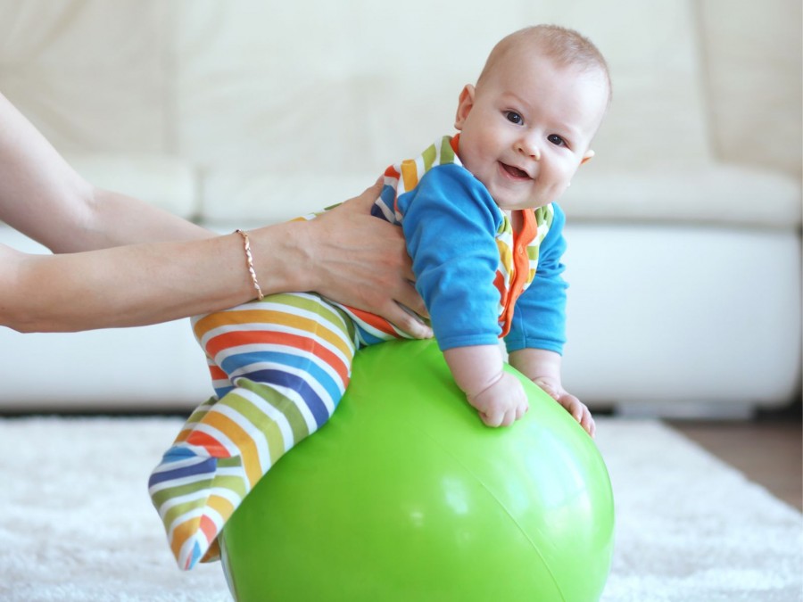 Ein lachendes Baby in einem bunten Strampelanzug sitzt auf einem grünem Gymnastikball und wird von dessen Mutter festgehalten. - Gesundheitsamt - Canva von Alena Ozerova