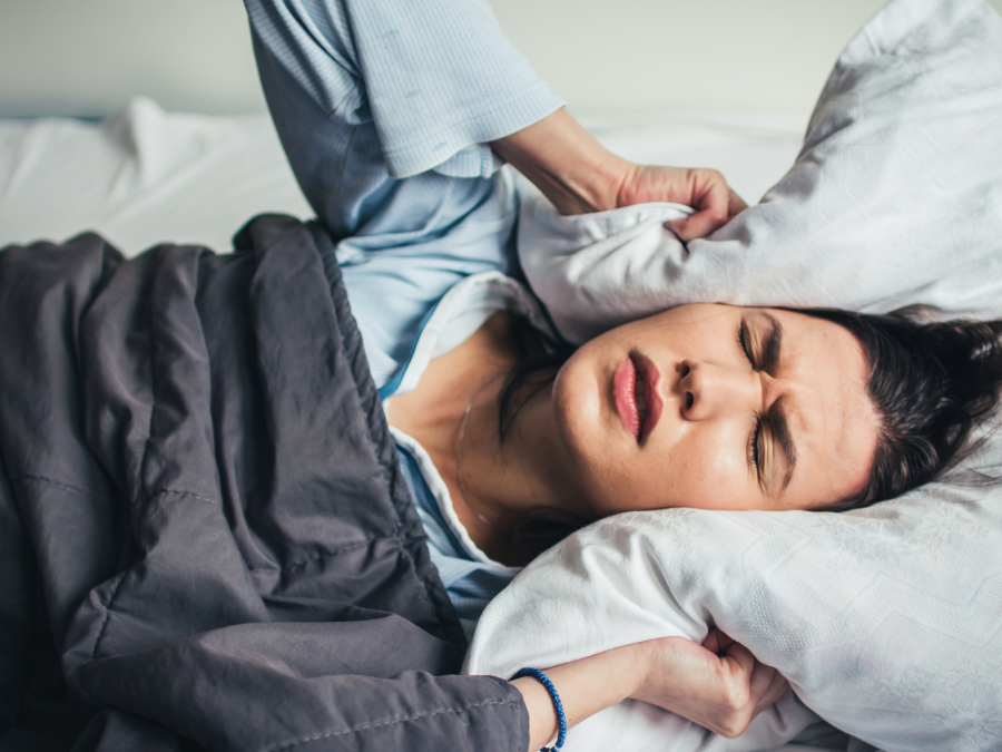 Frau im Bett drückt sich Kissen auf die Ohren und kann durch Lärm nicht schlafen. Lärmschutz, Ruhestörungen, Nachtruhe - Canva.com
