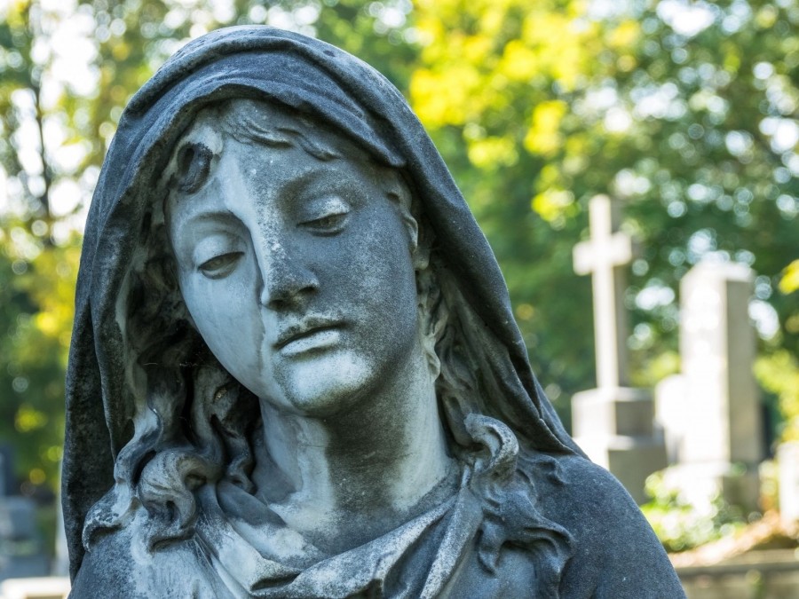 Trauernder Kopf einer weiblichen Statue auf einem Friedhof, einzelne Grabsteine im Hintergrund zu erkennen. Friedhöfe sind Orte der Erinnerung. - Canva