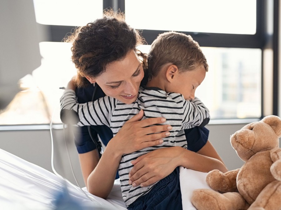 Eine Mutter umarmt ihren Sohn im Krankenhausbett. Rechts neben dem Kind sitzt ein brauner Teddy. - Gesundheitsamt - Canva von Rido