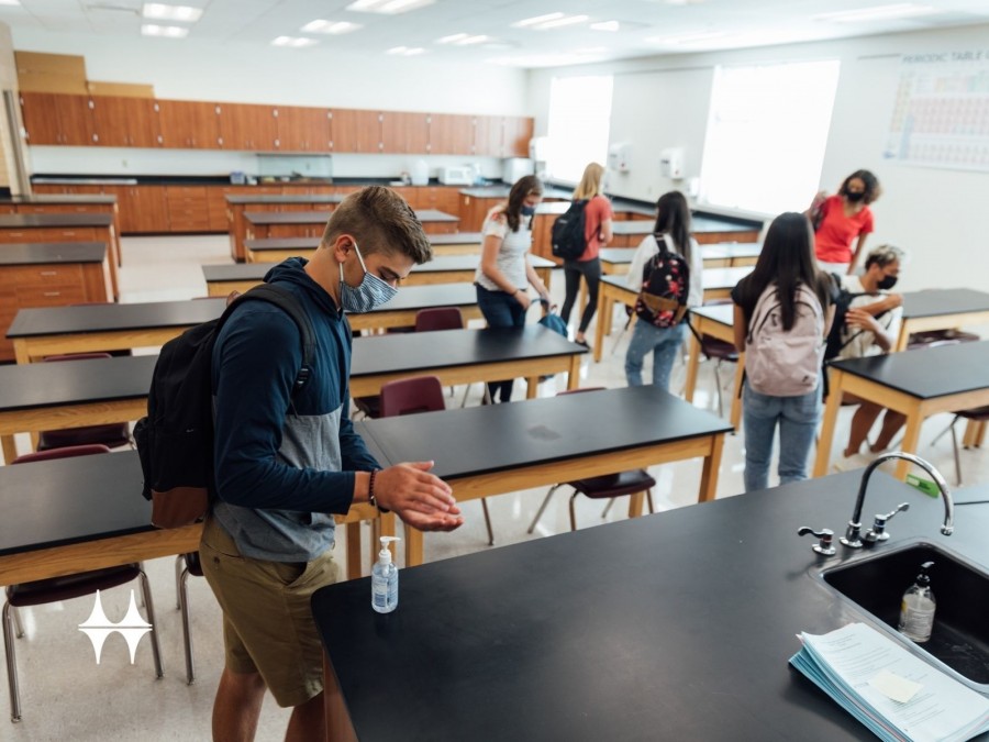 Schülerinnen und Schüler in einem Klassenraum. Ein Schüler desinfiziert sich seine Hände mit zur Verfügung gestellten Desinfektionsmitteln. Corona, Unterrichtsformen, Covid-19 - Canva
