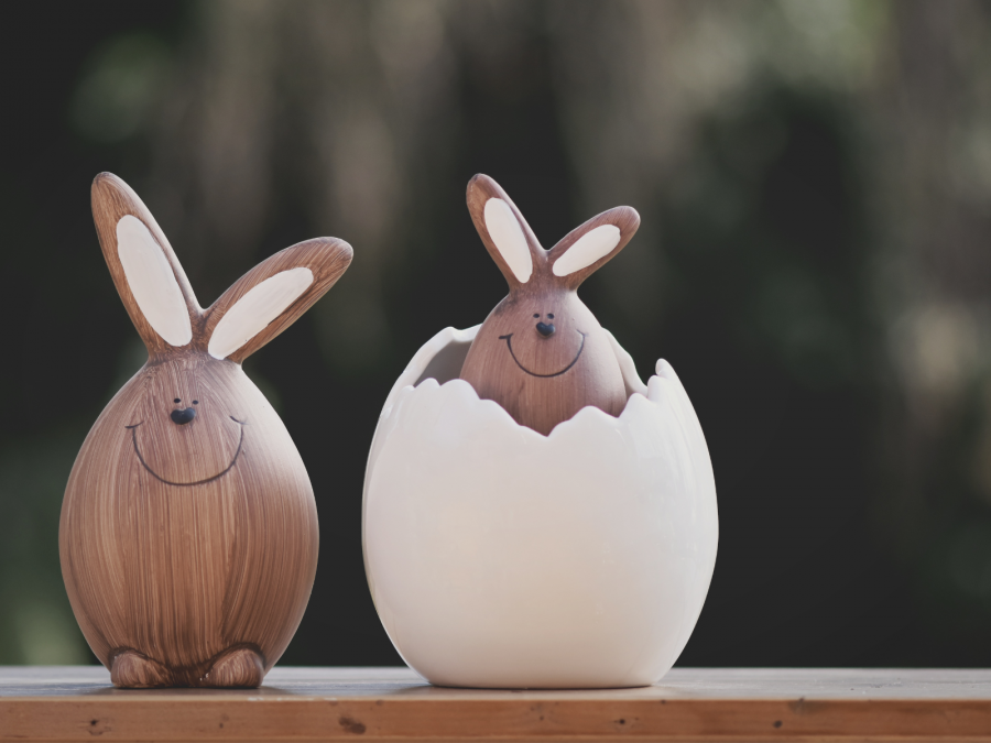 Osterhasendekoration: Ein brauner Keramikhase sitzt neben einem weißen, aufgebrochenen Keramikei. Aus dem Ei guckt ein kleinerer brauner Keramikhase. - Canva
