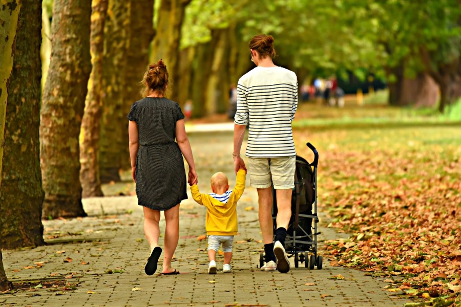 Foto zeigt junge Eltern beim Spaziergang im Park. Beide Eltern halten das Kind an den Händen, der Vater schiebt den Kinderwagen, Aufnahme von hinten.- MabelAmber auf Pixabay