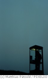 Darstellung einer Person in einer Telefonzelle in abendlicher Dämmerung. Nachtaufnahme.