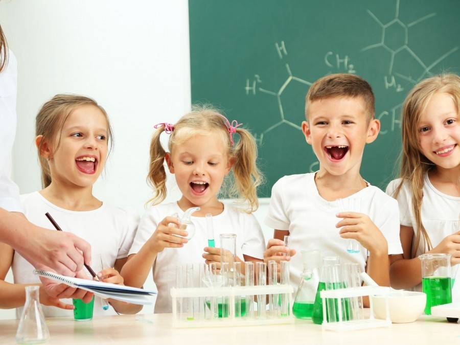 Das Foto zeigt eine Gruppe von Grundschulkindern, die im Chemieunterricht  biochemische Forschung betreibe. Im Hintergrund an einer Schultafel Chemieformeln, im Vordergrund Reagenzgläser mit grüner Flüssigkeit. - Canva