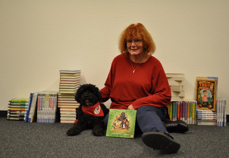 Birgit Hass zusammen mit ihrem Lesehund Rico auf dem Boden sitzend vor einer Reihe von Büchern - Stadtbibliothek/Birgit Hass