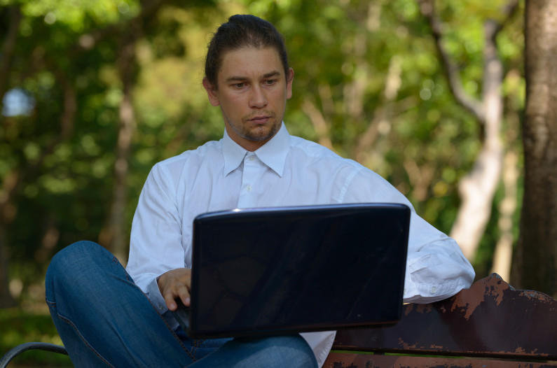 Ein Mann sitzt auf einer Parkbank und arbeitet am Notebook. - CANVA