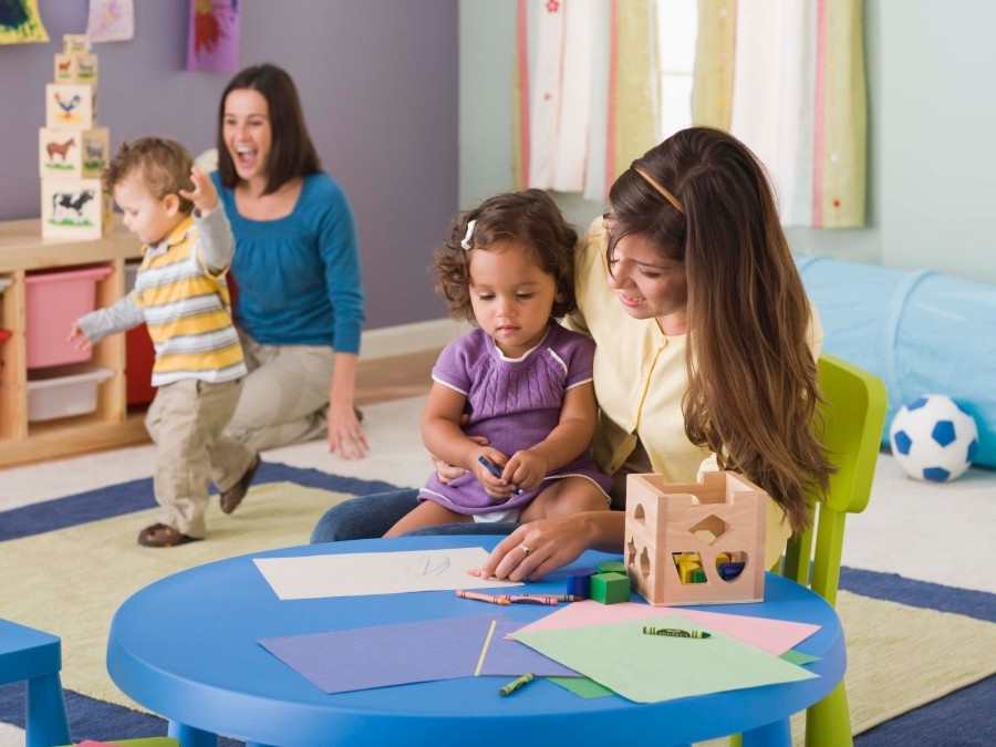 Zwei Kindertagespflegepersonen spielen mit Kleinkindern im Tagespflegenest. Kita, Kindergarten, Kinderbetreuung, Pflegevater, Pflegemutter - Canva