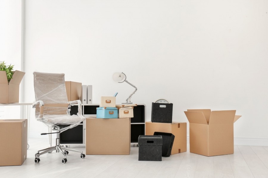 Raum mit Umzugskartons, einem noch eingepackten Bürostuhl und Schreibtischlampe. Symbolbild für Büromobilar. - Canva Raum mit Umzugskartons, einem noch eingepackten Bürostuhl und Schreibtischlampe. Symbolbild für Büromobilar. - Canva
