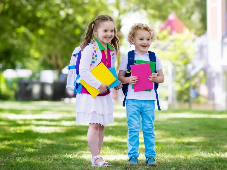 Junge und Mädchen stehen lachend vor der Schule an ihrem ersten Schultag mit bunten Heften in der Hand. - Gesundheitsamt - Canva von FamVeld