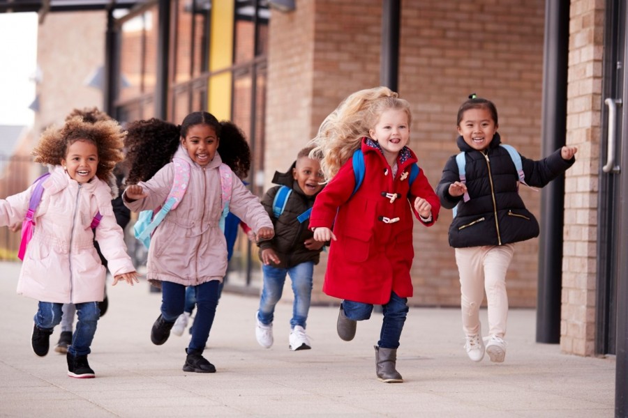 Mehrere Grundschulkinder rennen mit ihren Schultaschen auf dem Rücken lachend an der Schule vorbei. Grundschulen, Einschulung, - Canva