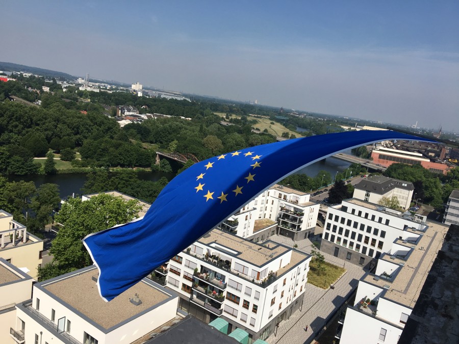 Das Foto zeigt die Europaflagge am Rathausturm in Großaufnahme. Im Hintergrund ein Panoramabild mit Blick auf die Gebäude an der Ruhrpromenade. - Onlineteam