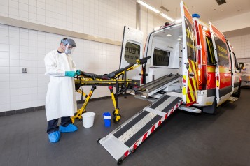 Das Bild zeigt eine Person mit Schutzkleidung in einer Halle, die eine Krankentrage eines Krankentransportwagen desinfiziert. - Feuerwehr Mülheim an der Ruhr