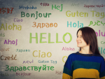 Junge Frau steht vor einer Tafel auf der in bunten Farben und vielen Sprachen Begrüßungen geschrieben sind. Übersetzende, Sprachkundigenpool, Dolmetschende - Canva