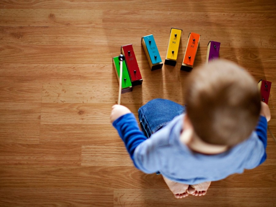Kleinkind hockt auf dem Boden und spielt mit einem Glockenspiel. Kindertagespflege - Canva