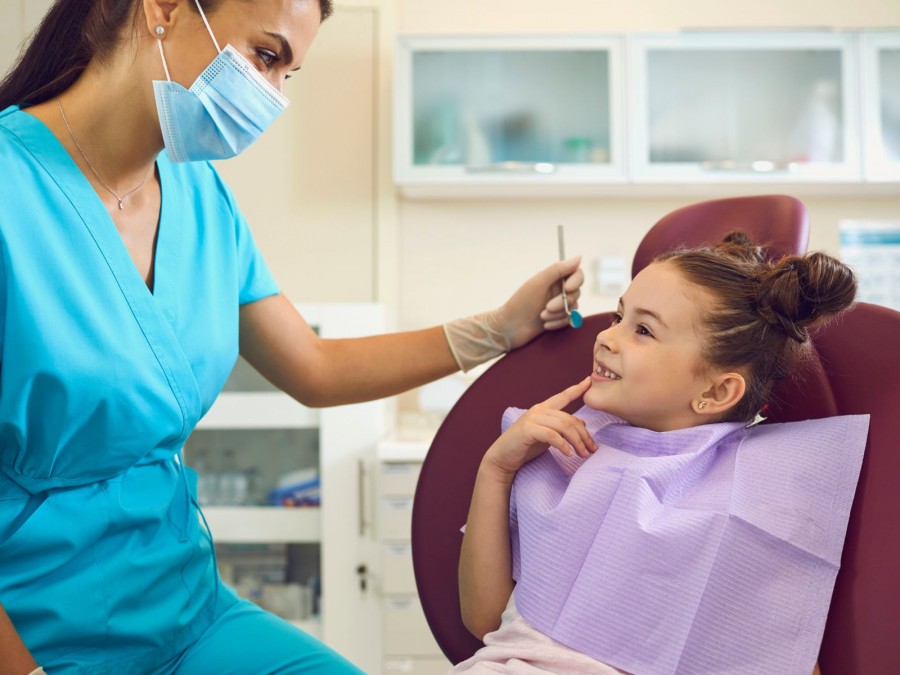 Ein junges Mädchen mit braunen Haaren und zwei Zöpfen sitzt lächelnd in einem Behandlungsstuhl in einer Praxis für Zahnmedizin. Gegenüber sitzt eine Zahnärztin in einer blauen Praxisbekleidung und hält in der linken Hand einen Mundspiegel. - Gesundheitsamt - Canva von Studioroman