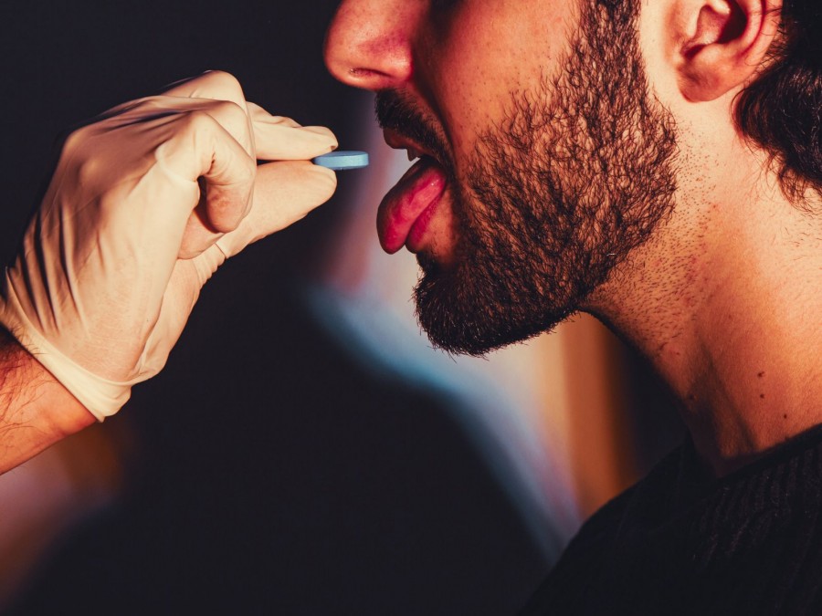 Mann mit Bart öffnet den Mund um eine blaue Pille einzunehmen die ihm von einer Hand mit einem  Schutzhandschuh entgegengehalten wird. - Gesundheitsamt - Canva von Corelens