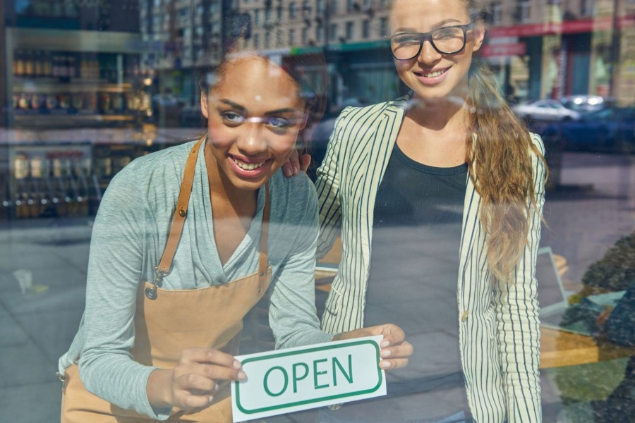 Zwei Frauen stehen lächelnd hinter der Schaufensterscheibe ihres neu eröffneten Geschäftes und befestigen ein OPEN-Schild. Neueröffnung, Gewerbe - Canva