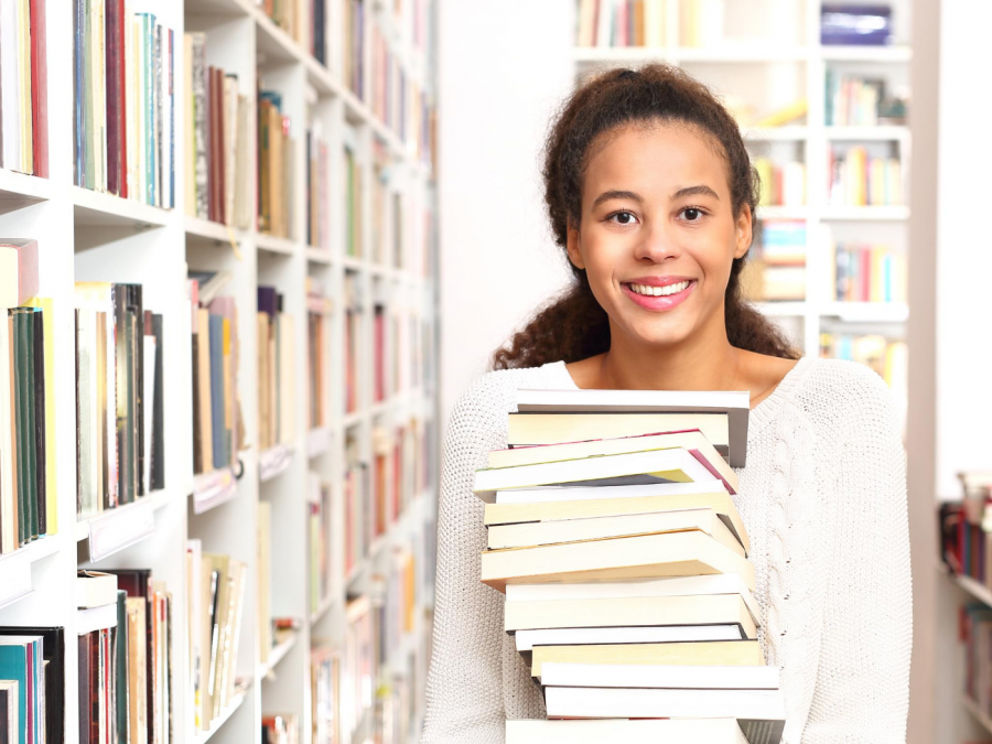 Junge Mitarbeiterin in der Bibliothek, sie trägt zwischen Bücherregalen einen Stapel Bücher und lacht. Ausbildung, Fachangestellte für Mediendienste und Informationsdienste - Canva