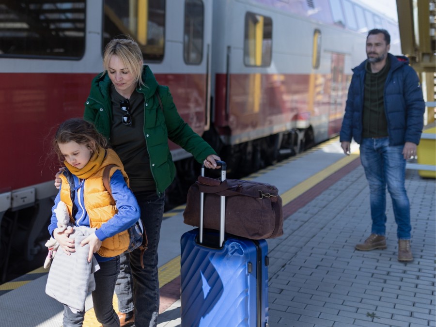 Das Foto zeigt eine Frau mit einem Mädchen, welches mit Koffern und Reisetasche auf einem Bahnsteig stehen. Im Hintergrund ist ein Zug zu sehen und ein Mann schaut Ihnen nach. - Online Redaktion - Referat I - Canva - halfpoint