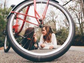 Zwei junge Frauen, auf der Straße liegend und lachend, durch ein Fahrrad-Rad und Speichen fotografiert. - Quelle/Autor: Photo by Seth Doyle on Unsplash