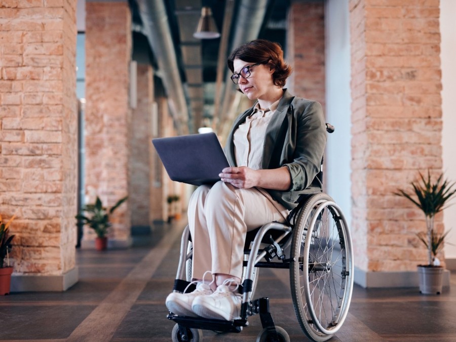 Frau im Rollstuhl, sie schaut in ihren aufgeklappten Laptop auf ihrem Schoß. Barrierefreiheit, Behinderungen, Schwerbehindertengesetz - Canva