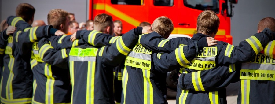 Rückansicht: Team von mehreren Feuerwehrmännern, die Arm in Arm eine Reihe bilden, vor einem Einsatzfahrzeug. Zusammenhalt, Feuerwehrleute, Teamarbeit - Berufsfeuerwehr Mülheim an der Ruhr