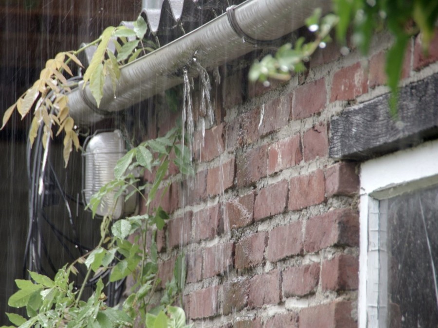 Regenrinne mit kleinen Löchern an einem Häuschen, es regnet so stark, dass die Regenrinne überläuft. Niederschlagswasser, Versickerung - Canva