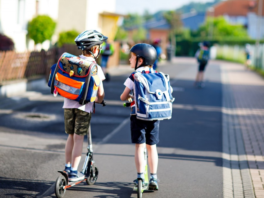 Schulweg, Sicherheit, Verkehrswacht, Schulkinder, Roller: Das Foto zeigt zwei Schuljungen mit Tornistern auf ihren Rollern auf der Straße in einer Wohnsiedlung. - Canva