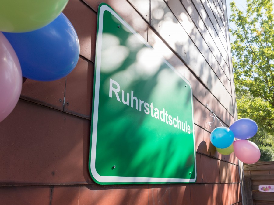 Das neue Schild an der Ruhrstadtschule wurde feierlich geschmückt. Drei bunte Luftballons hängen links und rechts vom Schild. - Online Team, Referat I - Helena Grebe