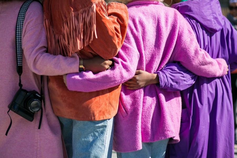 Von hinten fotografiert: Vier Frauen in knallbunten Mänteln halten sich in den Armen, Arm in Arm. Symbol für Gleichstellungsstelle, Zusammenhalt - Photo by Vonecia Carswell on Unsplash