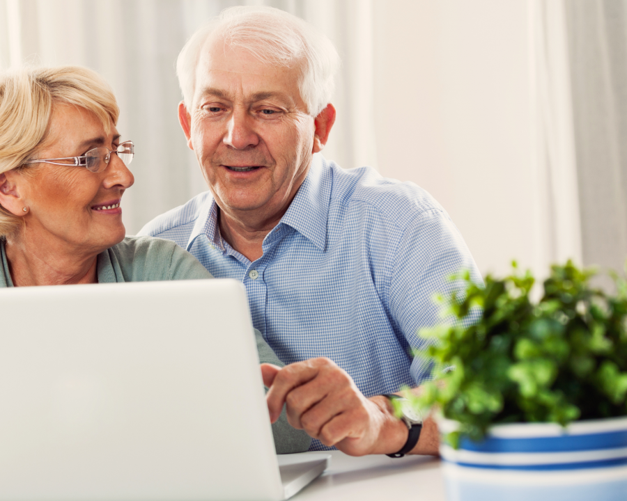 Ein älterer Mann und eine ältere Frau sitzen zusammen vor einem Laptop und scheinen sich über das zu unterhalten, was sie daran gemeinsam tun. - Amt 19 / CANVA
