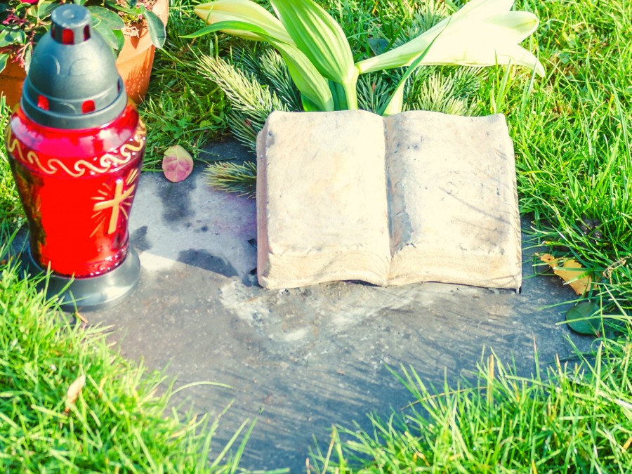 Friedhof, Friedhöfe, Brandgefahr: Das Foto zeigt ein rotes Grablicht auf einem auf dem Boden liegenden Grabstein auf der eine Lilie liegt. - Canva