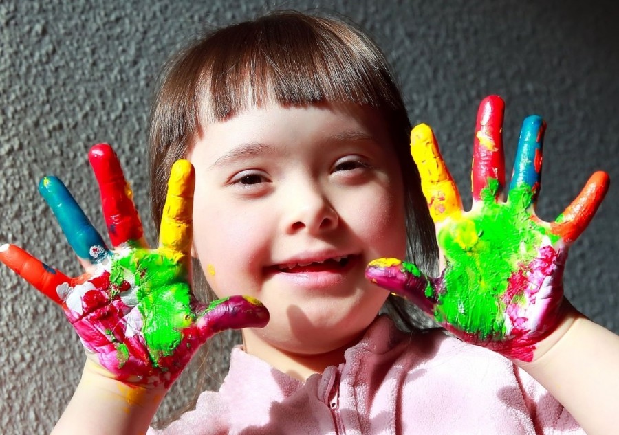 Kleines Mädchen hält ihre bunt bemalten Hände hoch und grinst. Inklusion, Behinderung, Beeinträchtigung, Tagespflegenest, Kita, Kindergarten, Kinderbetreuung, Pflegevater, Pflegemutter, Kindertagespflege - Canva