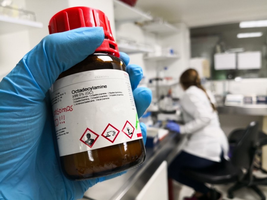 Hand mit blauem Gummihandschuh in einem Labor hält eine Flasche mit einer Chemikalie auf der Gafahrstoff-Symbole aufgeklebt sind. - Canva