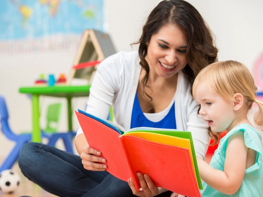 Braunhaarige Frau zeigt im Kindergarten einem kleinen Mädchen mit Zöpfen ein buntes Buch. - Canva von SDI Prductions