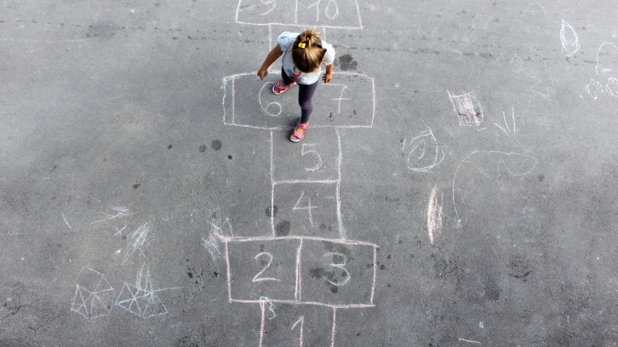 Ein kleines Mädchen spielt auf einem Schulhof Hüpfkästchen - Canva
