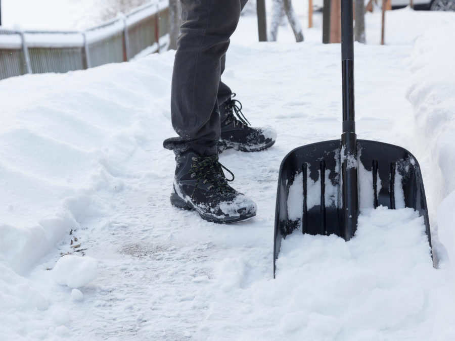 Eine Schaufel und Beine einer Person im Schnee, die Wege von Schnee befreit. Winterdienst, Straßenreinigung - Canva