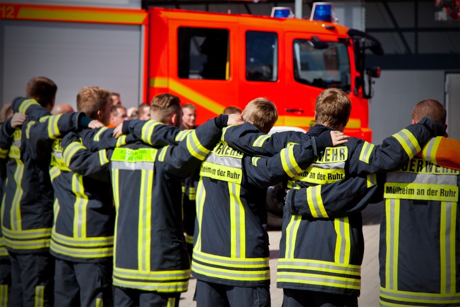 Rückenansicht: Team von mehreren Feuerwehrmännern, die Arm in Arm eine Reihe bilden, vor einem Einsatzfahrzeug. Zusammenhalt - Berufsfeuerwehr Mülheim an der Ruhr