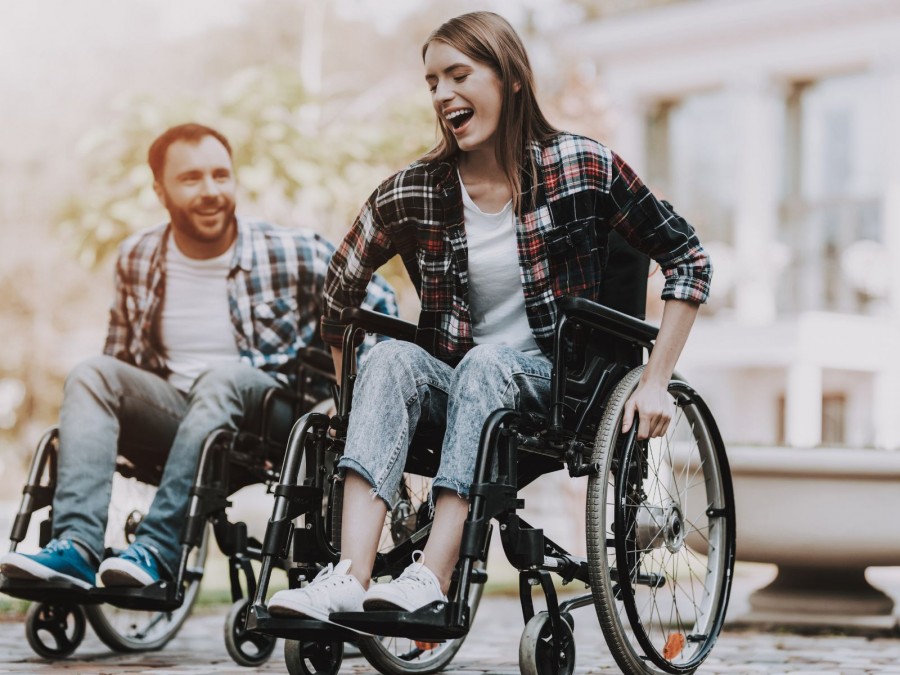 Ein lachender Mann und eine lachende Frau fahren in ihrem Rollstuhl durch einen Park. Beide sin in Jeans, Karohemd und Sneaker gekleidet. - Gesundheitsamt, Canva - von vadimguzhva
