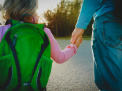 Bildausschnitt mit einem Elternteil, der sein kleines Kind mit grünem Rucksack an der Hand hält und aus dem Bild laufen. Tagespflegenest, Kita, Kindergarten, Kinderbetreuung, Pflegevater, Pflegemutter, Kindertagespflege