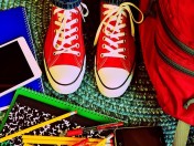 Rote Kinderturnschuhe auf einem Teppich, rundherum liegt Schulmaterial wie Stifte, Hefte, roter Schulrucksack und mehr. Schulwahl, Wechsel auf eine weiterführende allgemeinbildende Schule