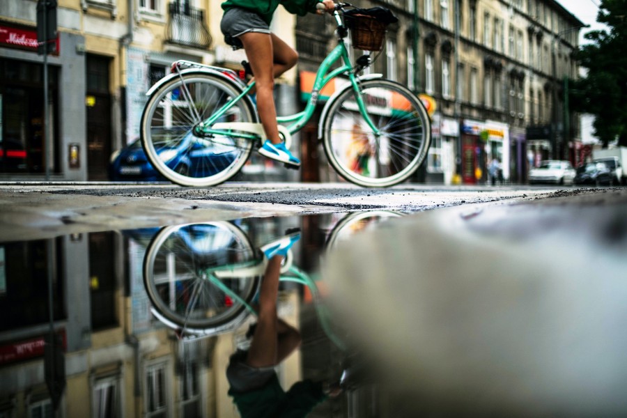 Frau fährt auf einem türkisen Fahrrad durch eine Straße, Bild spiegelt sich in einer Regenpfütze. - Photo by Erik Witsoe on Unsplash