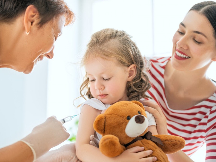 Eine Mutter hat Ihre Tochter auf dem Schoß. Das Mädchen hält einen Teddybären im Arm während eine Ärztin das Kind impft. - Gesundheitsamt - Canva von Yuganov Konstantin